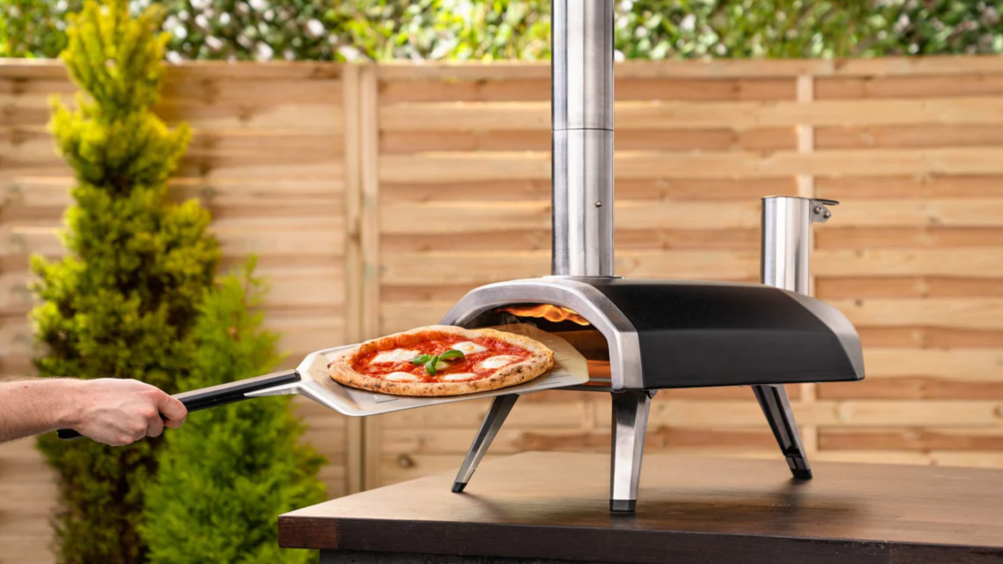 hand reaching for pizza from the ooni pizza oven, which is placed on a brown table
