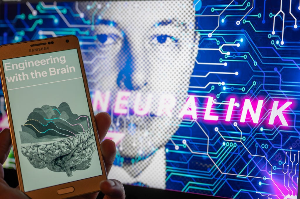 Neuralink website displayed on smartphone with founder Elon Musk seen on screen in the background.