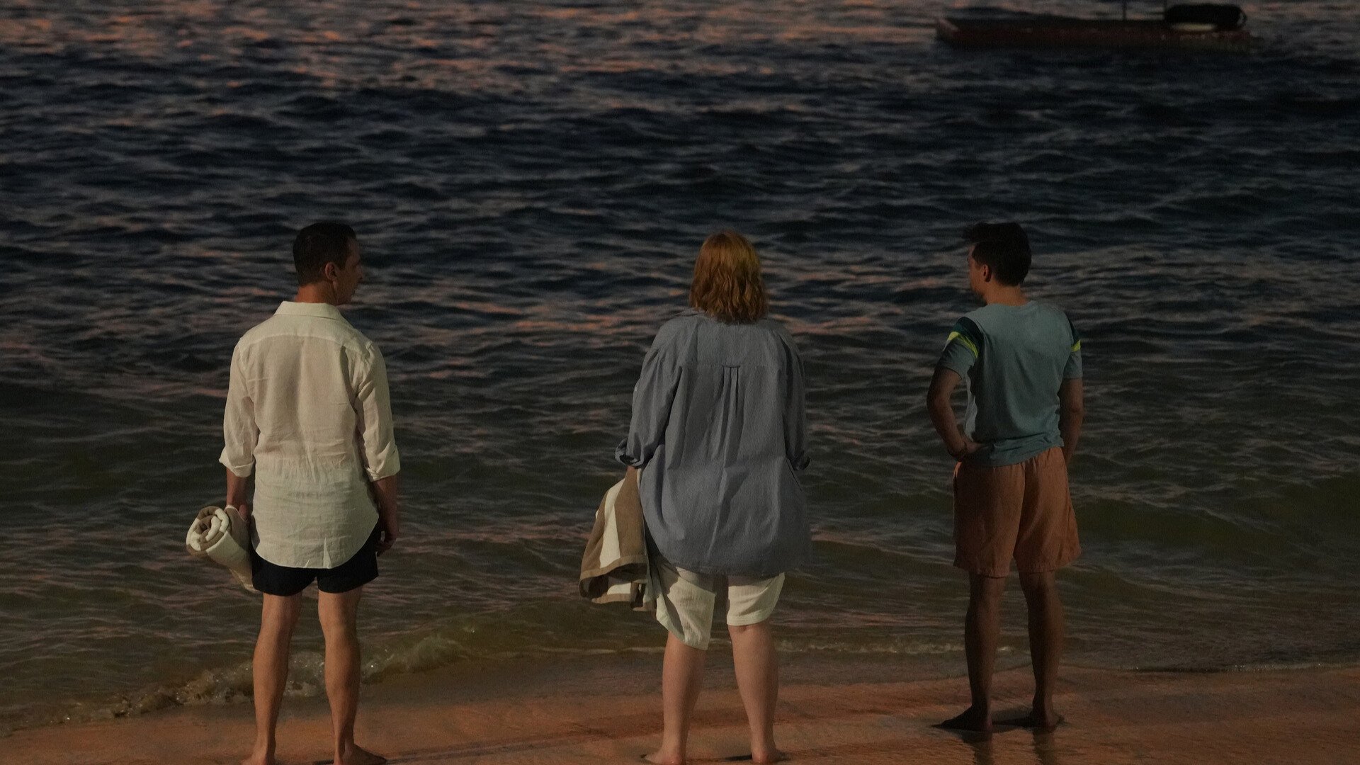 Three adults stand by a beach during sunset. 
