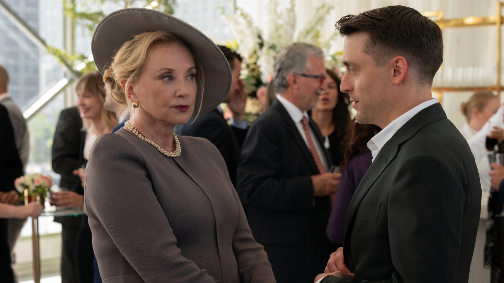A woman wearing a grey dress and hat, and a man wearing a suit, talk at an upscale function. 