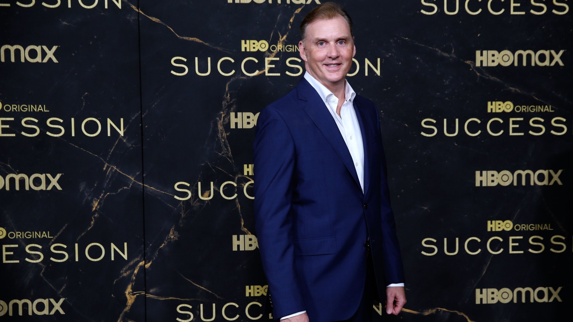 A man in a blue suit stands against a wall with the "Succession" logo repeated on it.