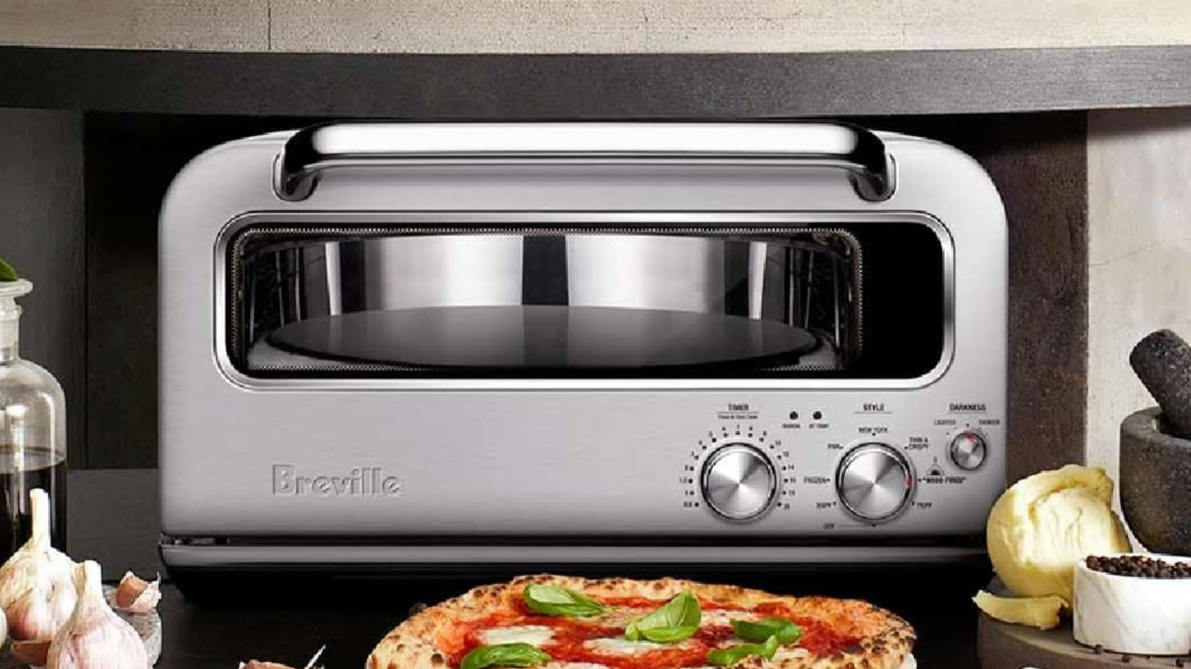 Breville pizza oven on kitchen counter with pizza in front, garlic to the left, mortar and pestle to the right