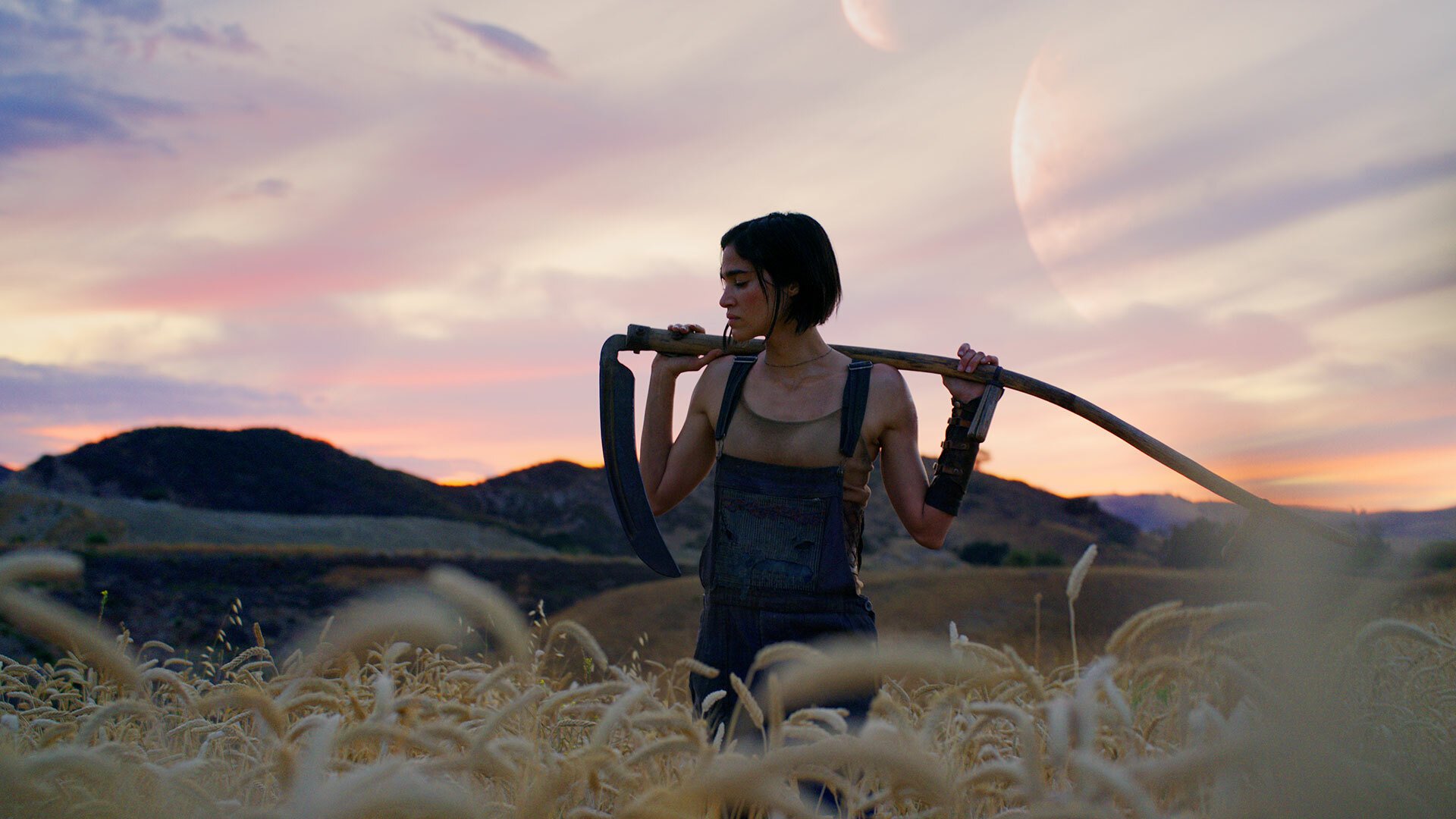 A woman holding a scythe stands in a corn field with two moons visible in the background.