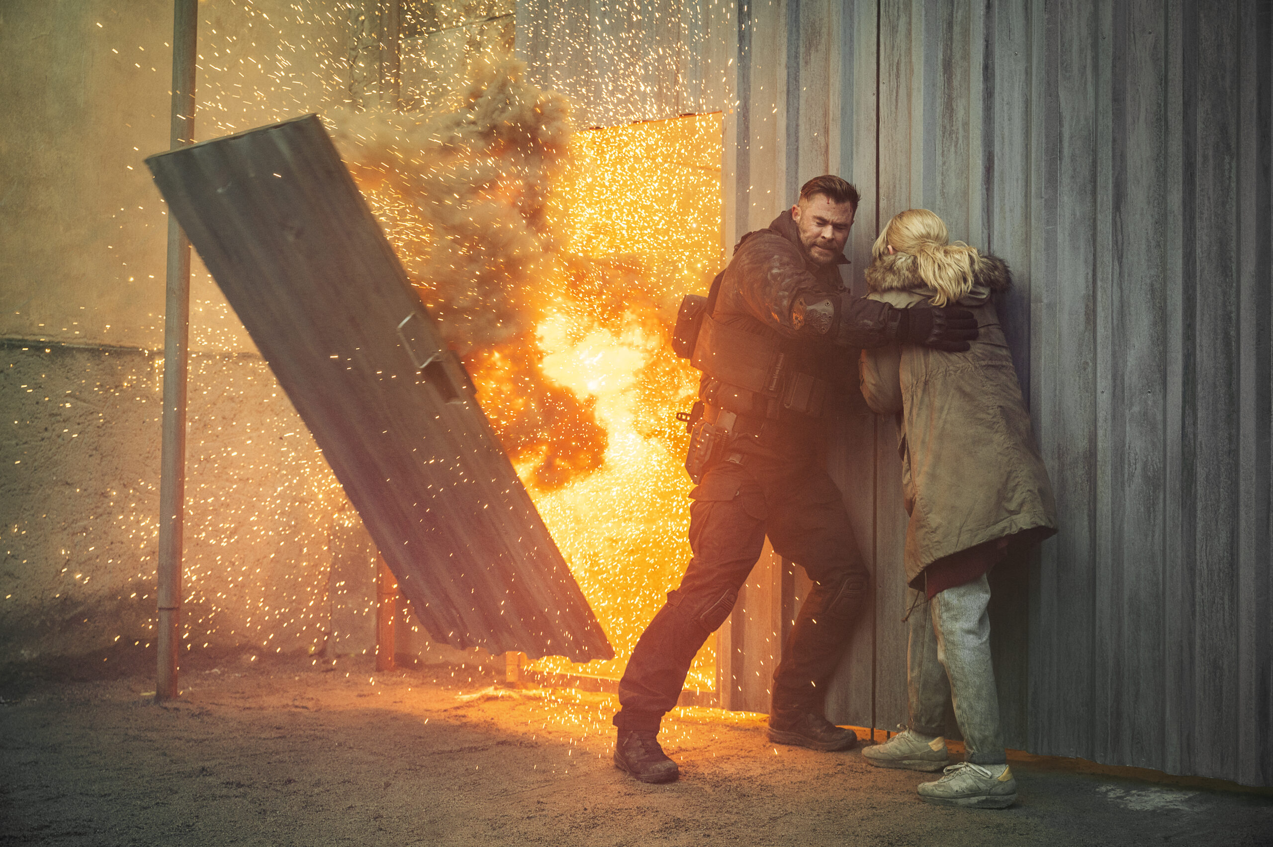 Chris Hemsworth stands to the side of an exploding building while shielding a woman.  