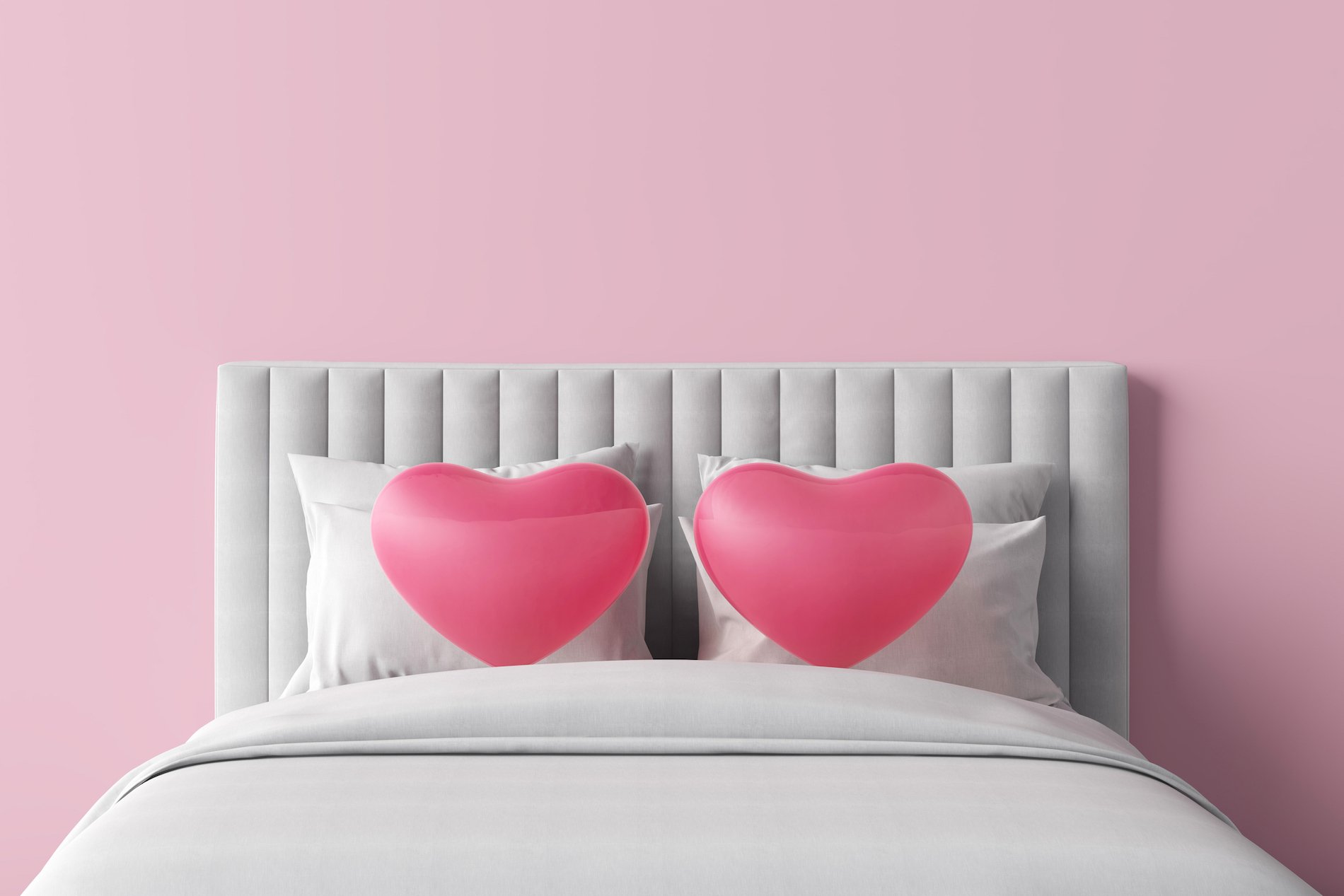 A bed with two heart shaped pink pillows, against a pink wall. 