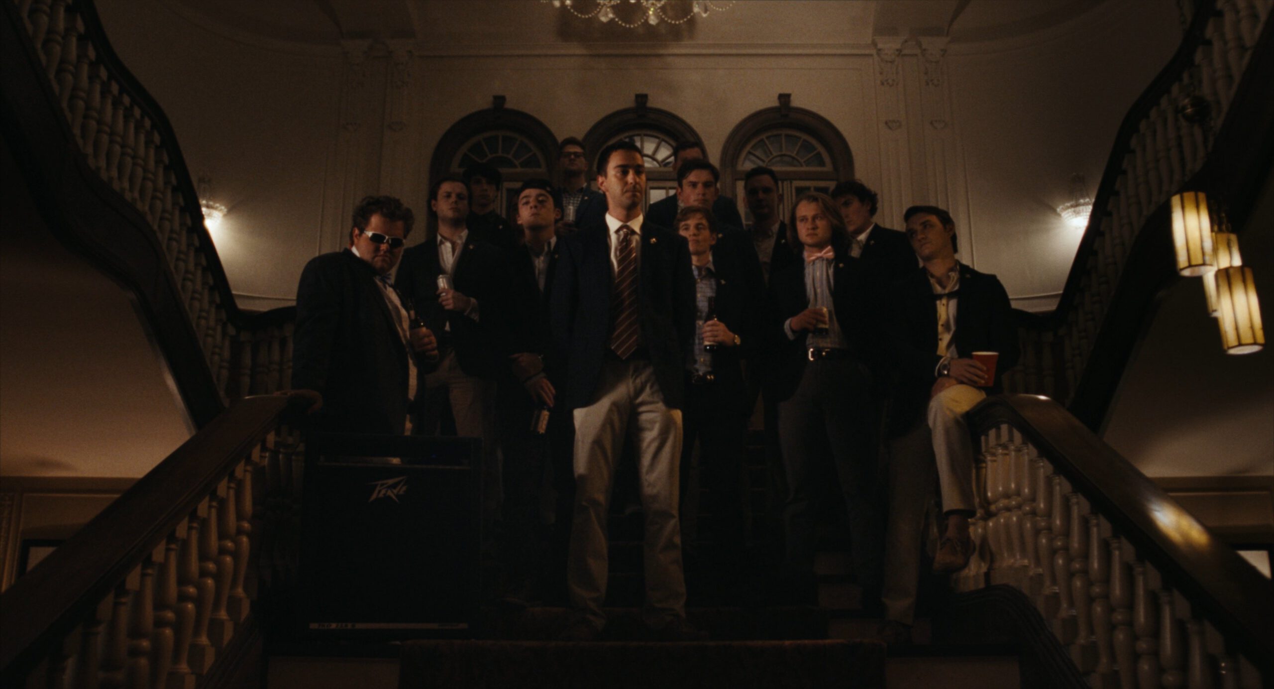 A group of frat brothers in suits stand on a grand staircase in a frat house.