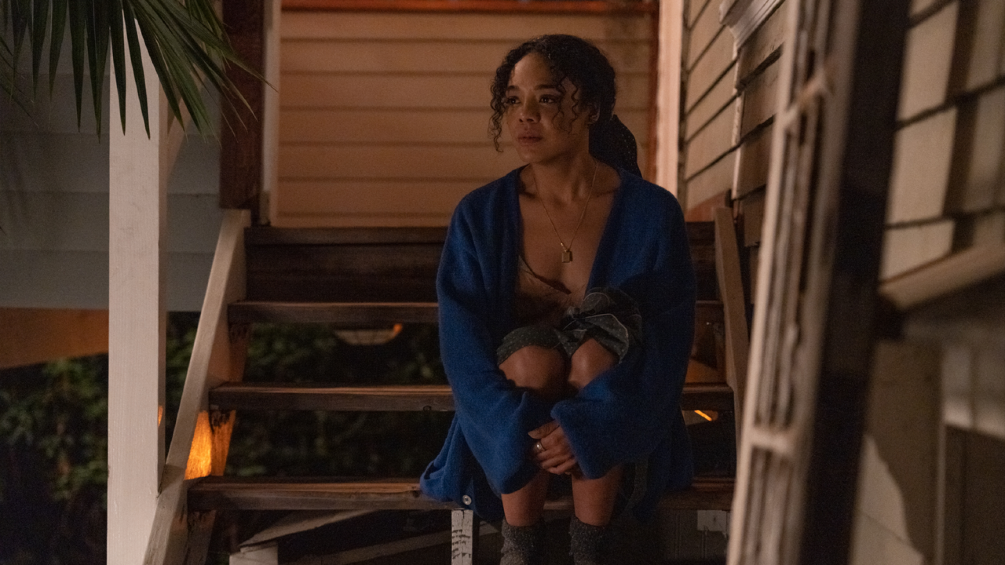 A woman in a blue sweater and pajamas sits on the stairs outside her house.