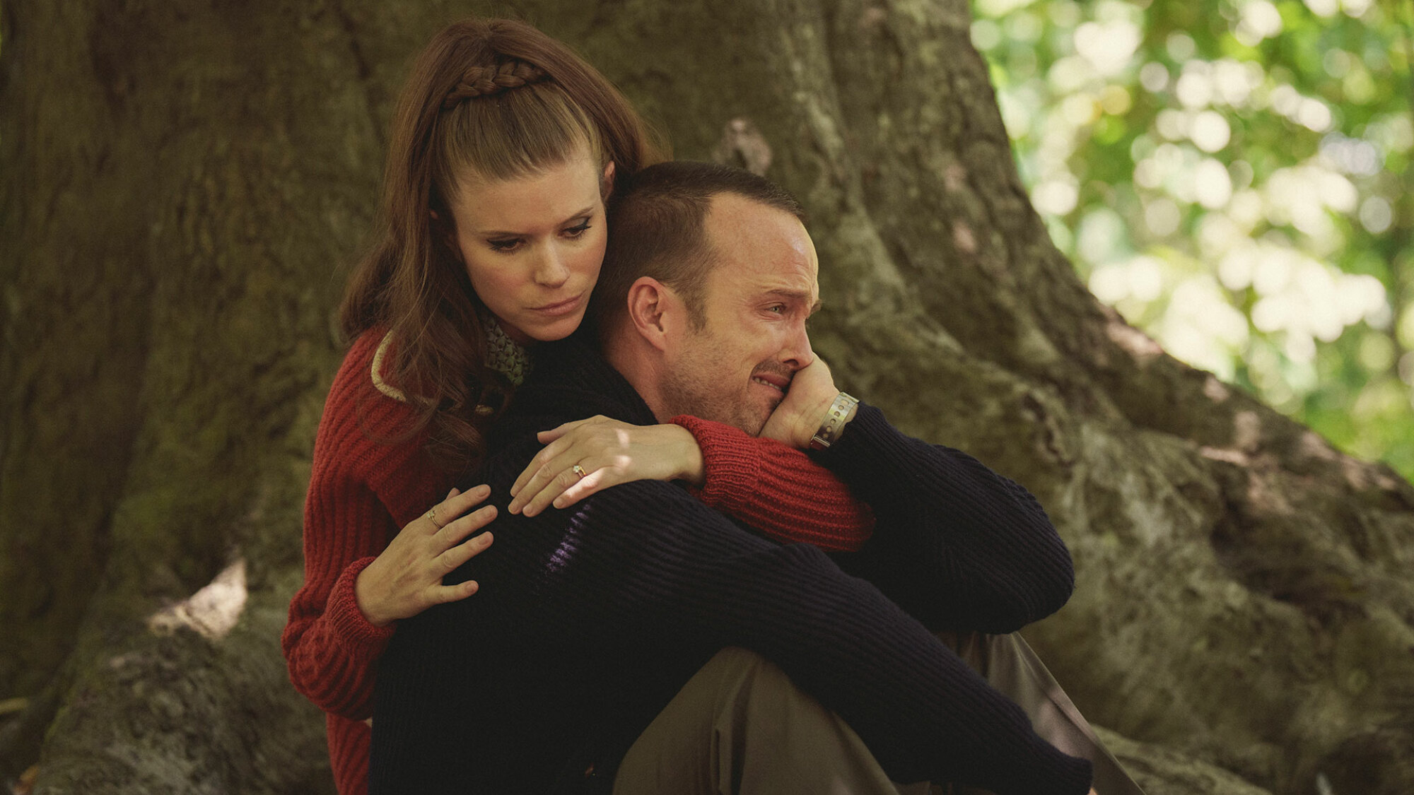A woman hugs a crying man at the foot of a tree.