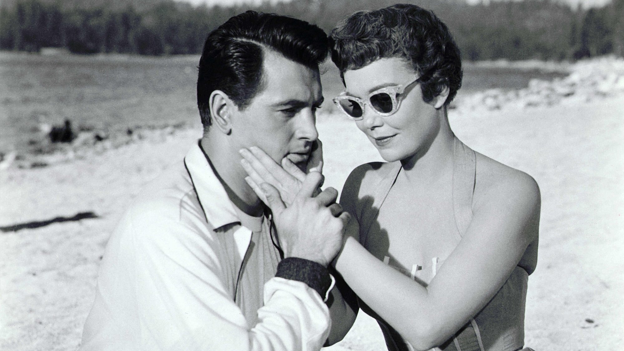A dark-haired man and a woman with short, dark hair sit on the beach together; her hand is on his cheek.