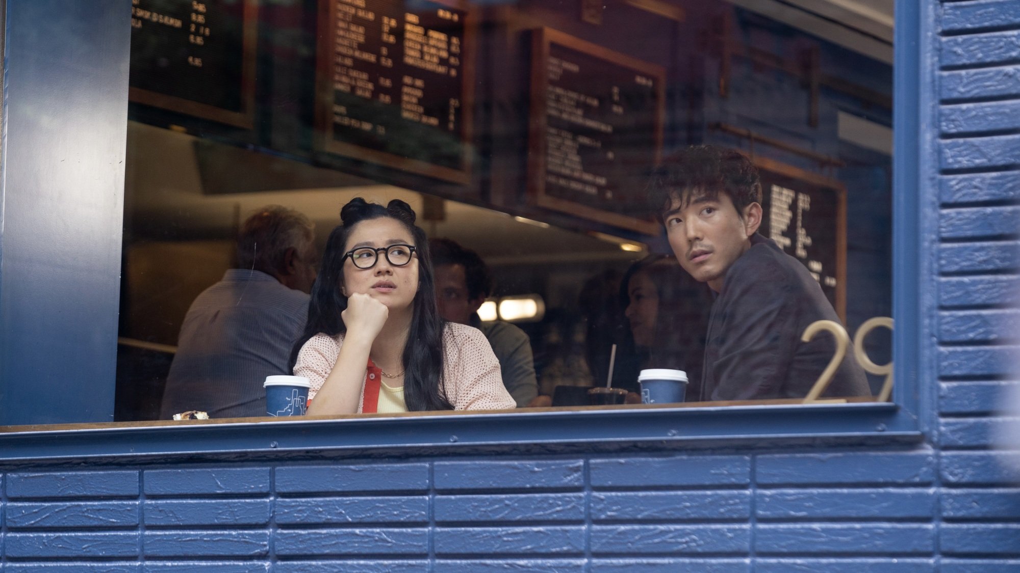 A man and woman look out the window of a coffee shop with a blue brick exterior.