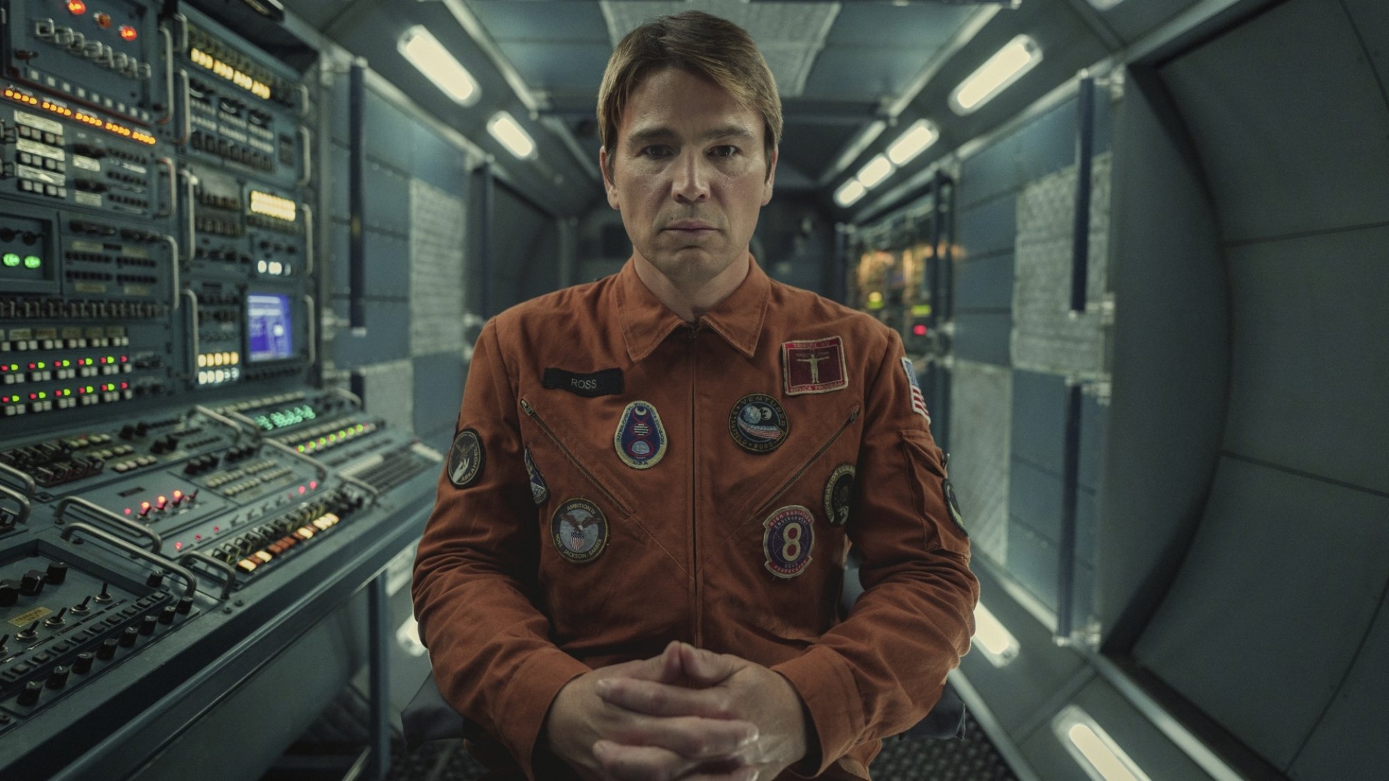 A man in an orange astronaut suit stares at the camera from inside a control room.