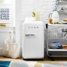 a white smeg fab 5 mini fridge in a kitchen next to a cart filled with dishware and a tote bag