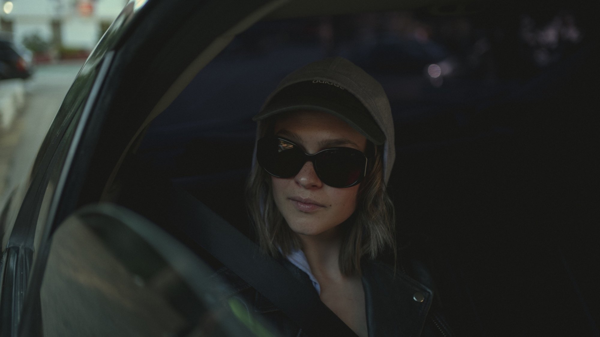 A woman in sunglasses and a hoodie sits in the back seat of a car with the window rolled down.