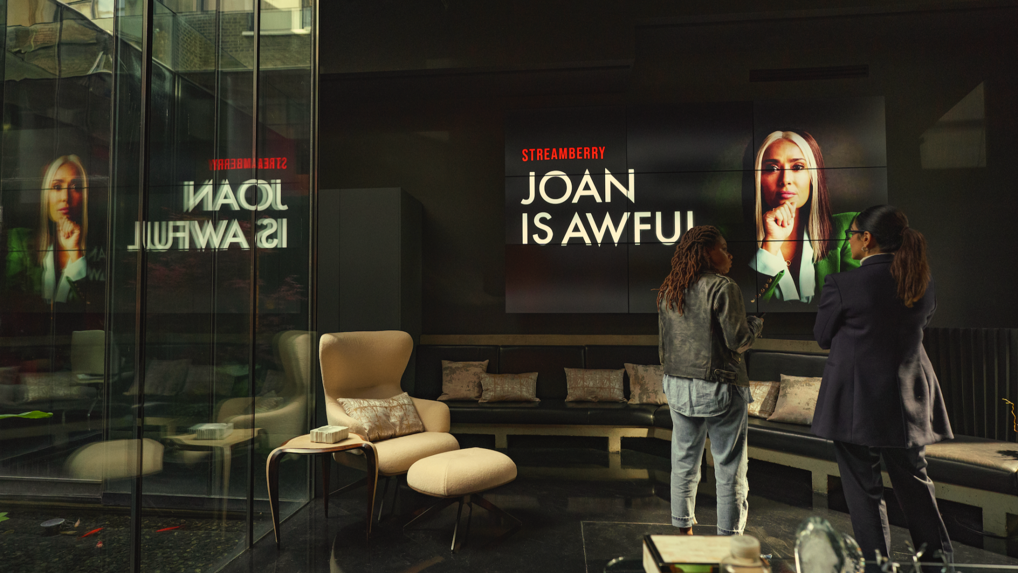 Two women stand in a conference room in front of a TV screen that says "Joan is Awful."
