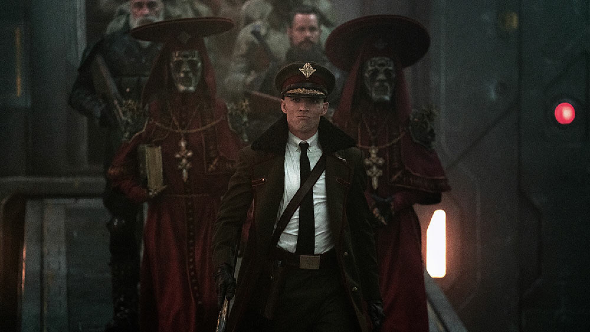 An armed man in uniform is followed by two lines of people, including a pair wearing red robes and face masks.