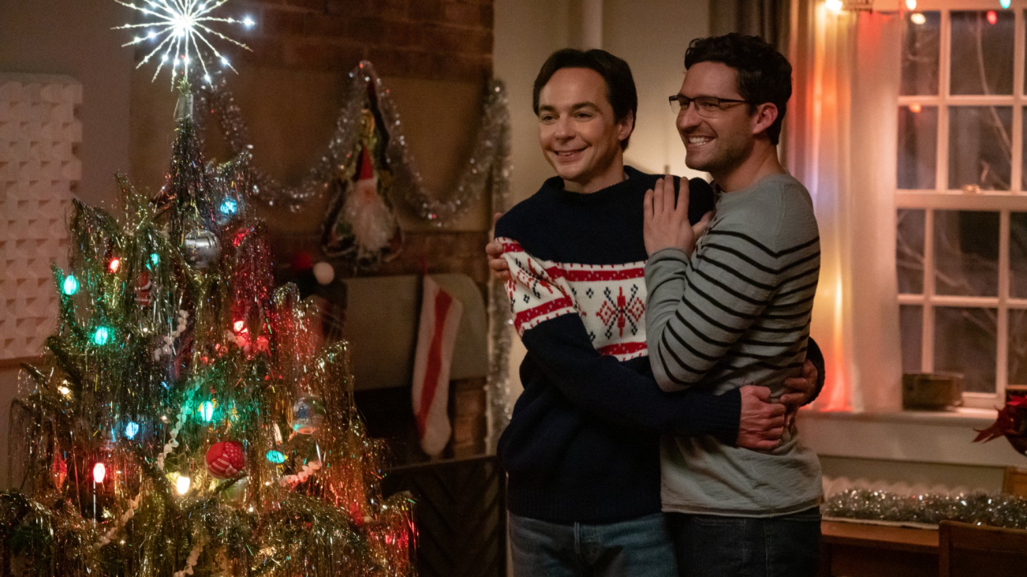 Two men embrace each other in front of a Christmas tree. 