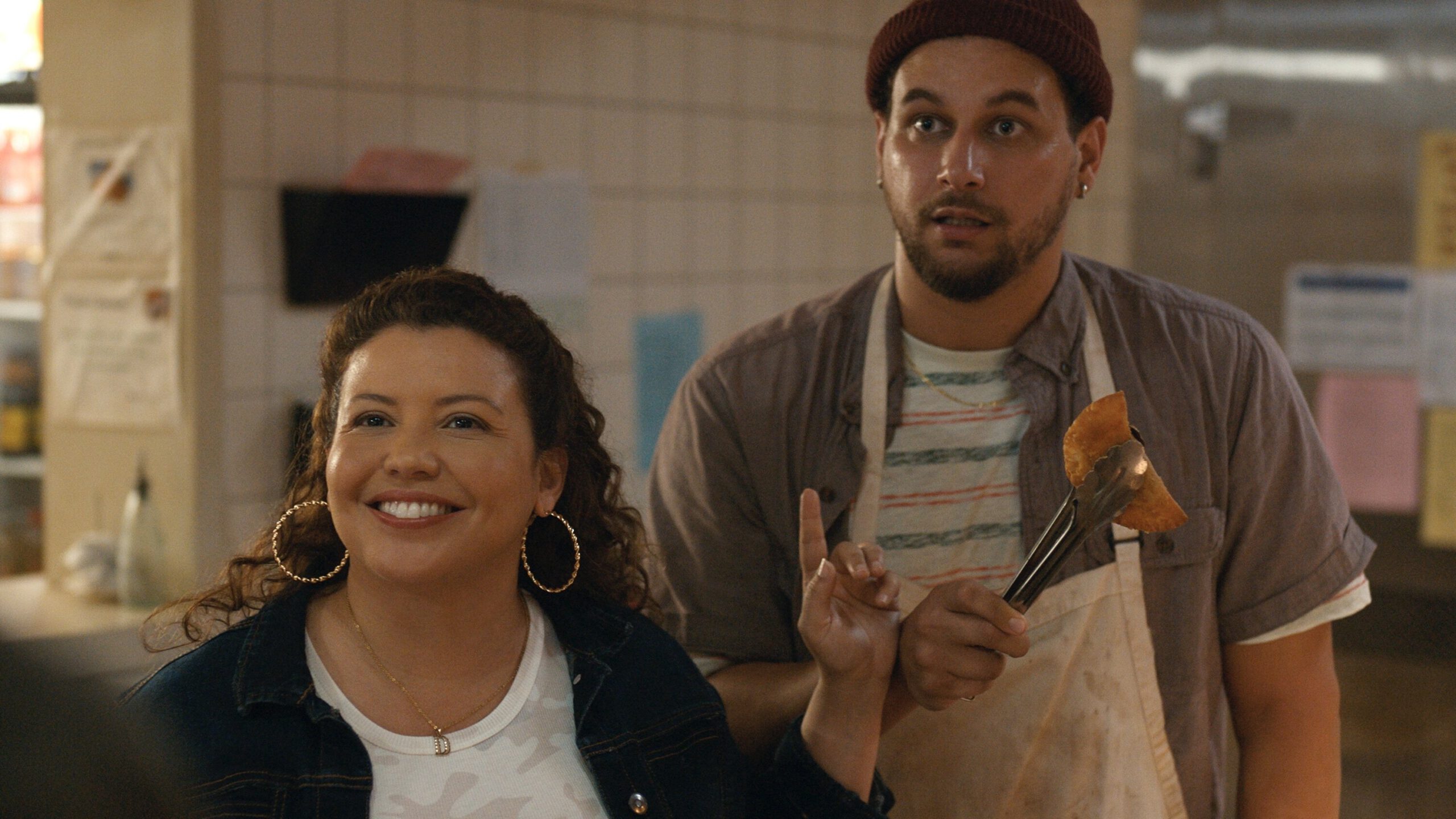 A woman in a white t-shirt and jean jacket smiles and points to a man in an apron and maroon beanie holding an empanada in some tongs.