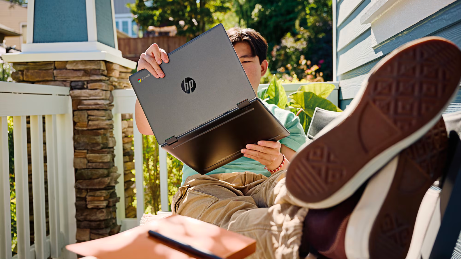 a teenage boy holding up an hp chromebook x360 and putting his legs up while sitting out on a sunny porch