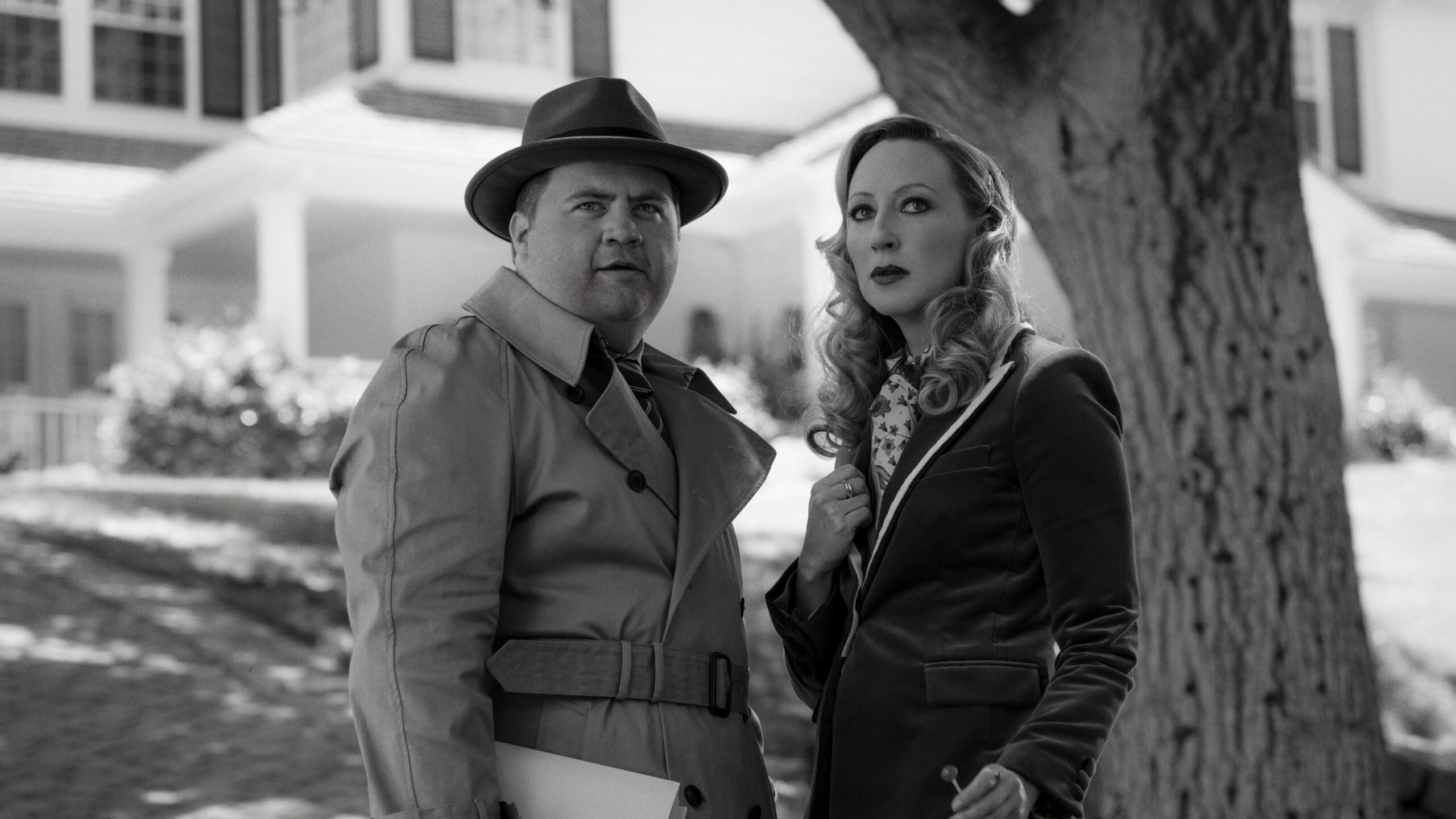 A man in a trenchcoat and fedora and a woman in a black suit stand together under a tree in a black and white film.