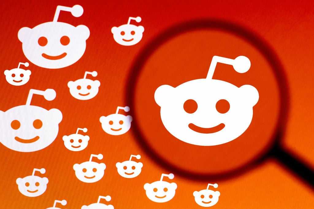 The Reddit logo seen displayed on a computer screen through a magnifying glass. A crowd of smaller Reddit logos is next to it.