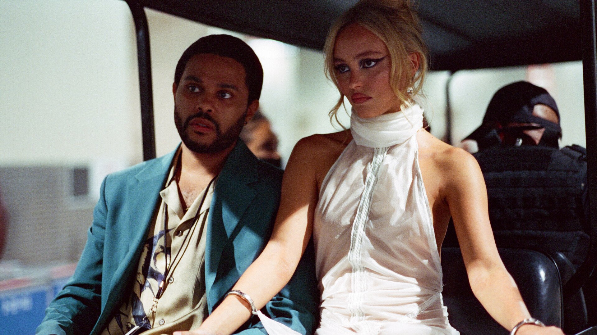 A man wearing a blue suit, and a woman wearing a white dress, sit in a golf cart inside a stadium's hallways. 
