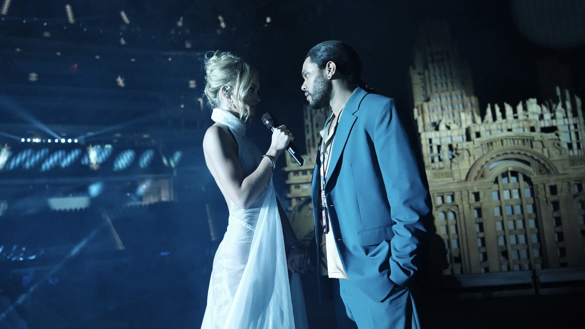 A woman in a white dress onstage at a stadium concert speaks to a man in a blue suit.