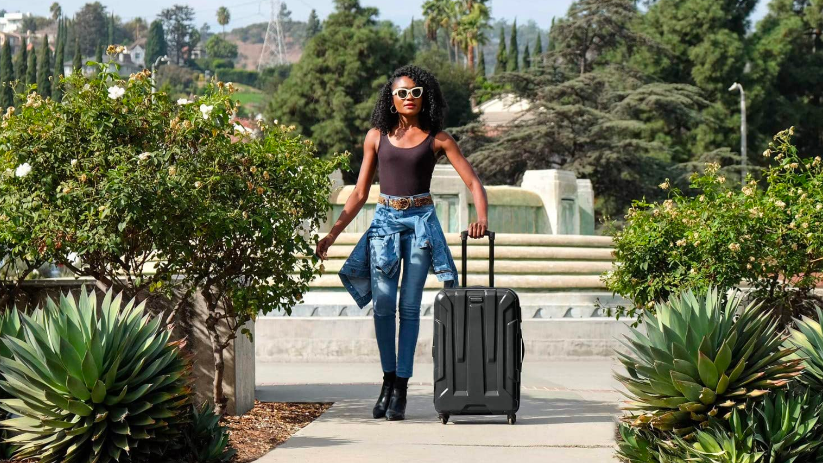 a woman in a brown tank top, jeans, and yellow sunglasses pulls a rolling samsonite suitcase through a garden on a sunny day