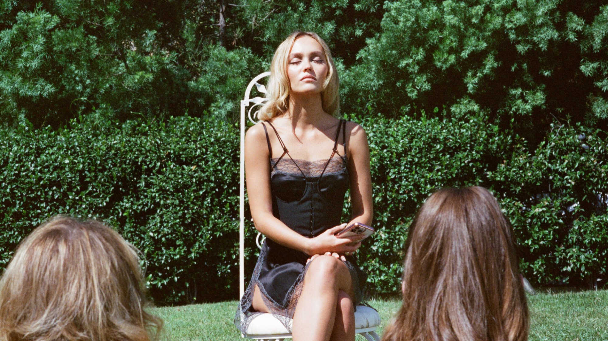 A woman wearing black lingerie rests on a white chair in a garden. 