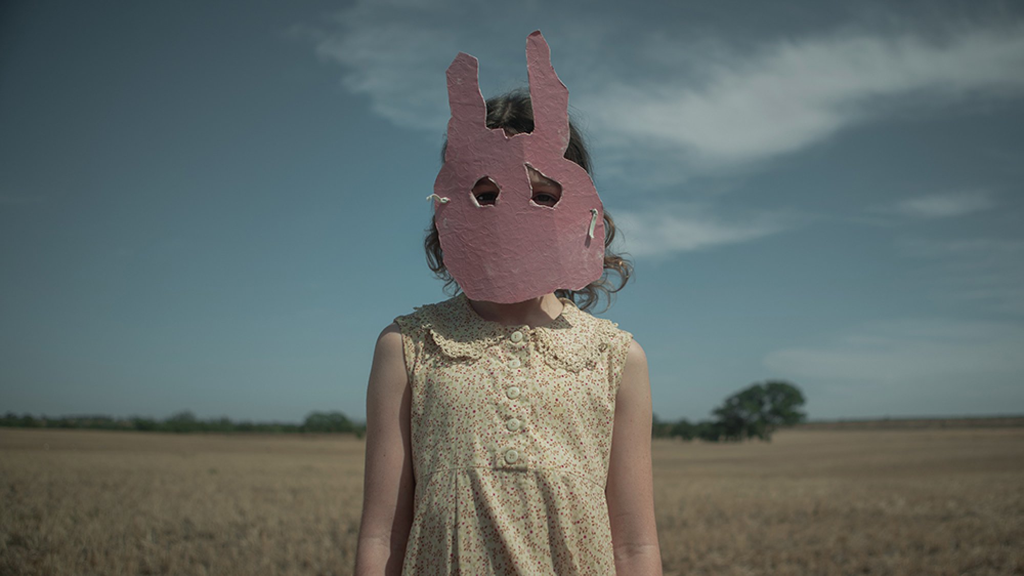 A girl in a pink rabbit mask stands in the wilderness.