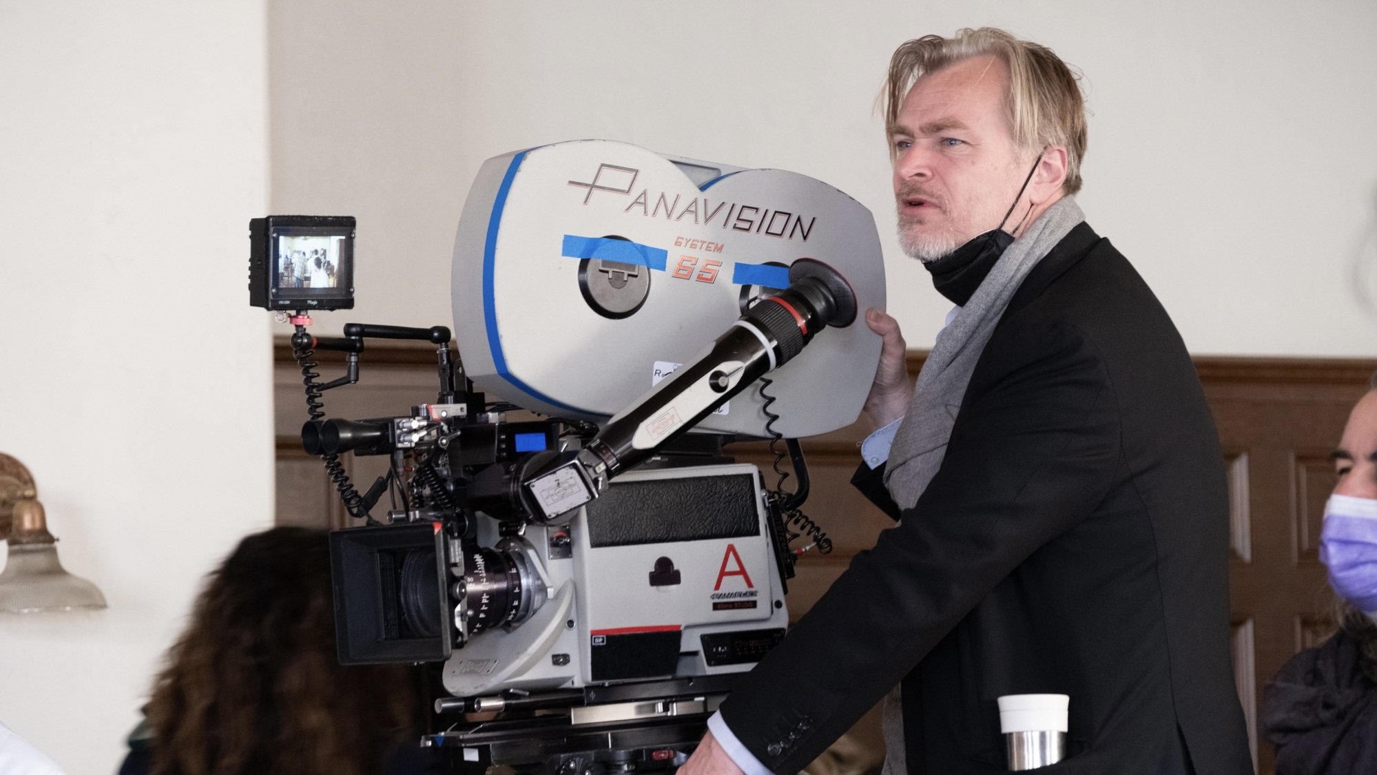 A man in a dark suit and grey scarf stands beside a large Panavision film camera.