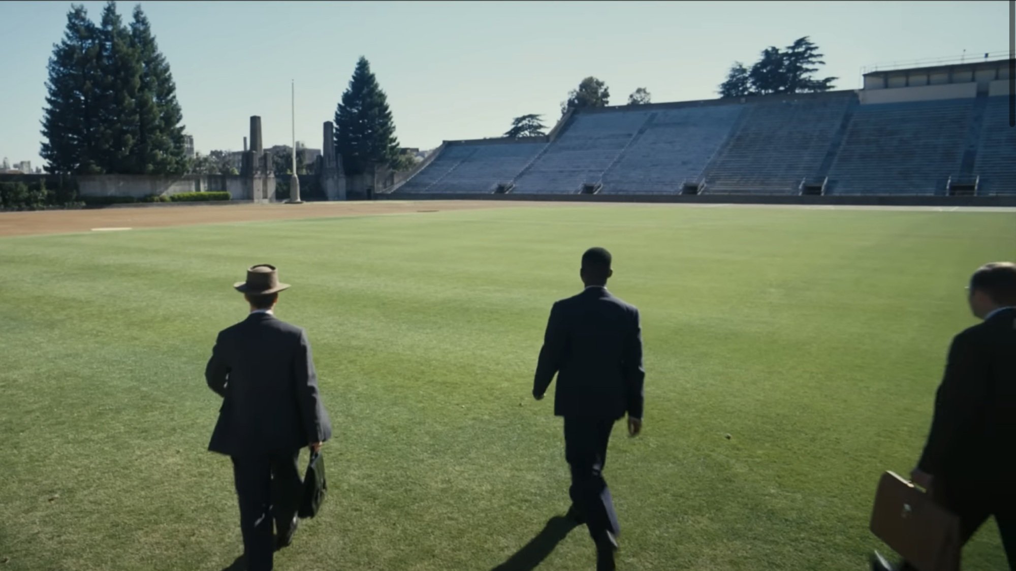 Three men in black suits walk across a college football field.