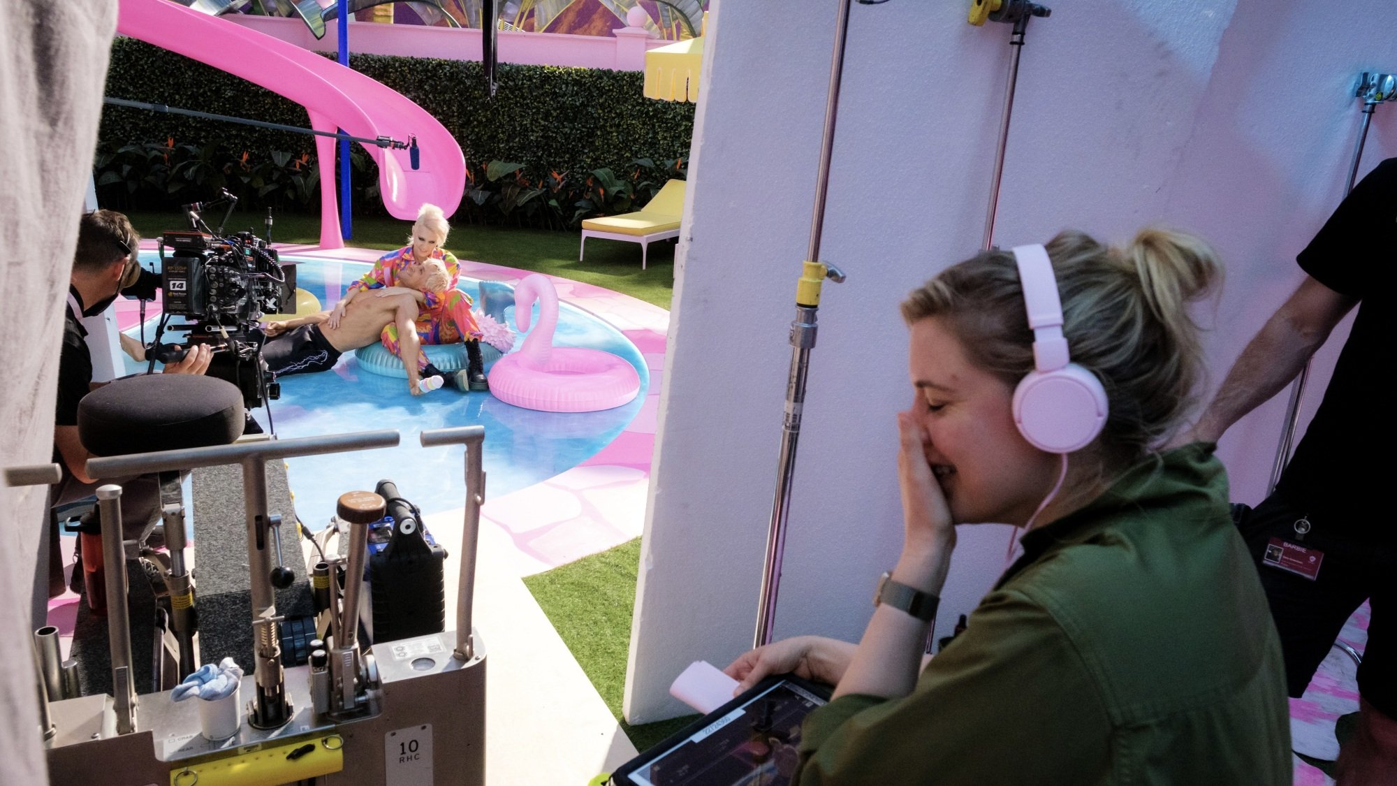 Greta Gerwig sits, laughing, as she directs a scene from "Barbie" where Ryan Gosling lies shirtless in Kate McKinnon's lap while they float on Barbie's fake pool.