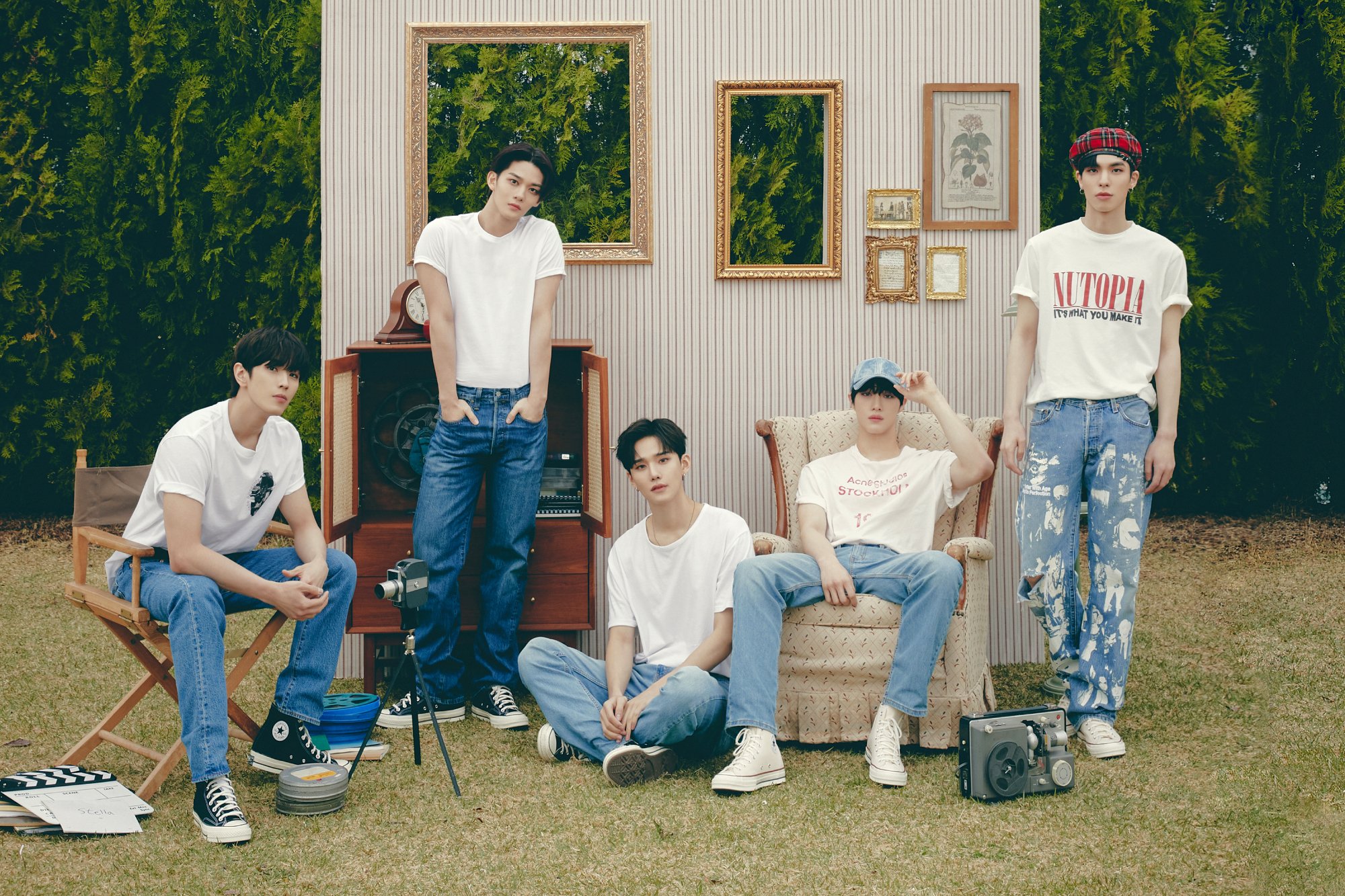 The band in white tees and light blue jeans, seated on a field with a faux living room wall behind them.