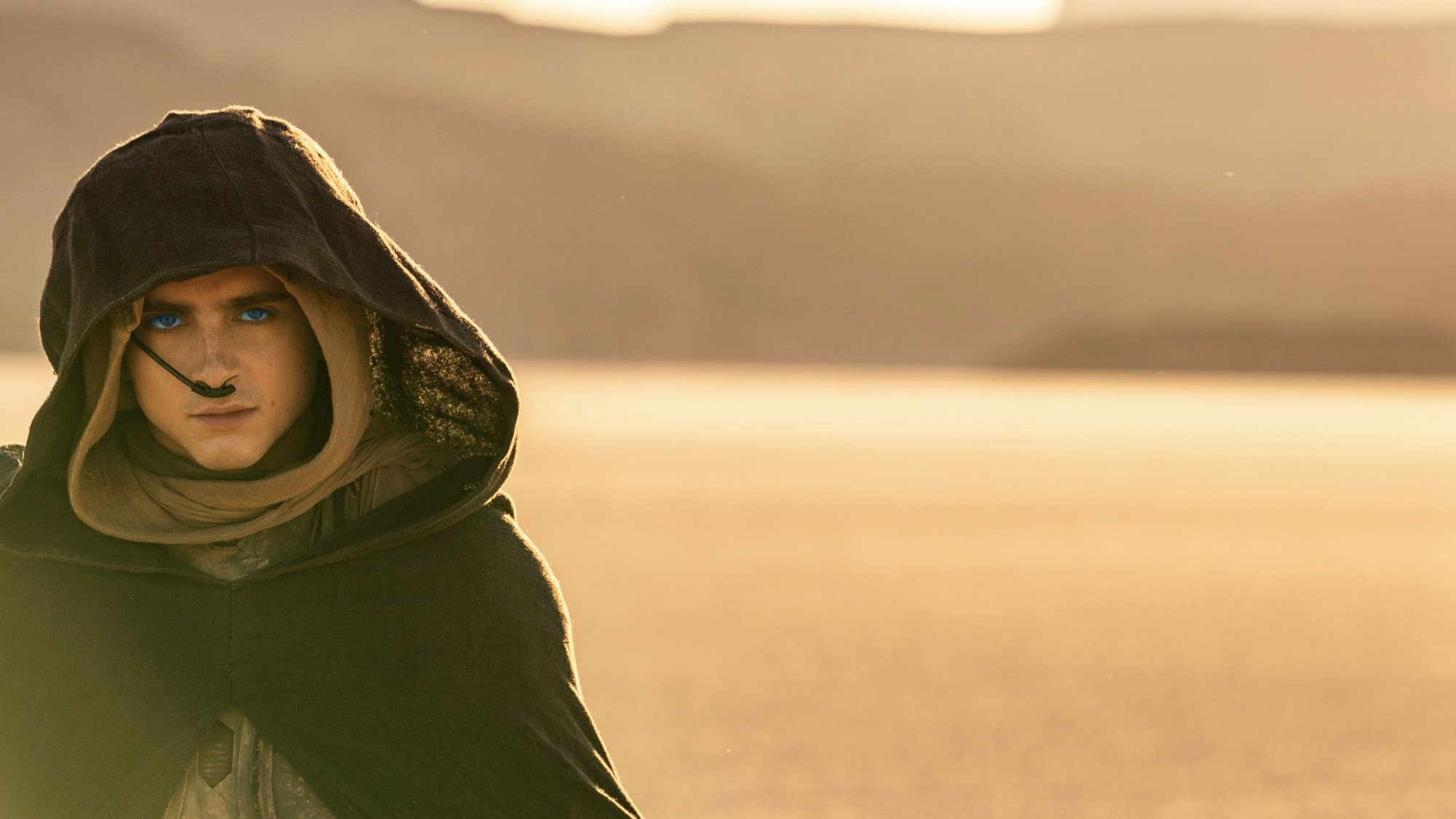 A young man with bright blue eyes walks through the desert in a dark, hooded cloak.