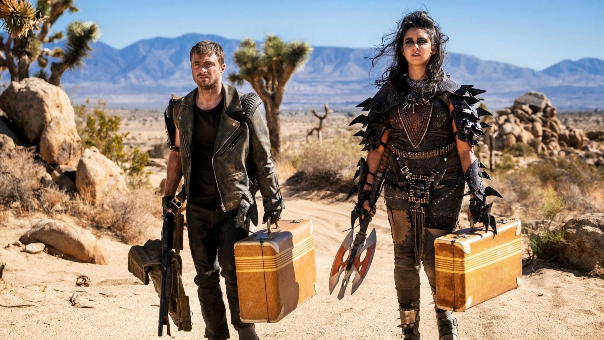 A man and a woman in biker outfits stand in a desert. 