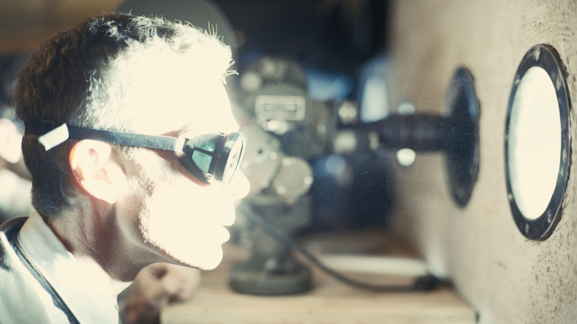A man in goggles stares out a circular window, bathed in brilliant white light from an explosion.