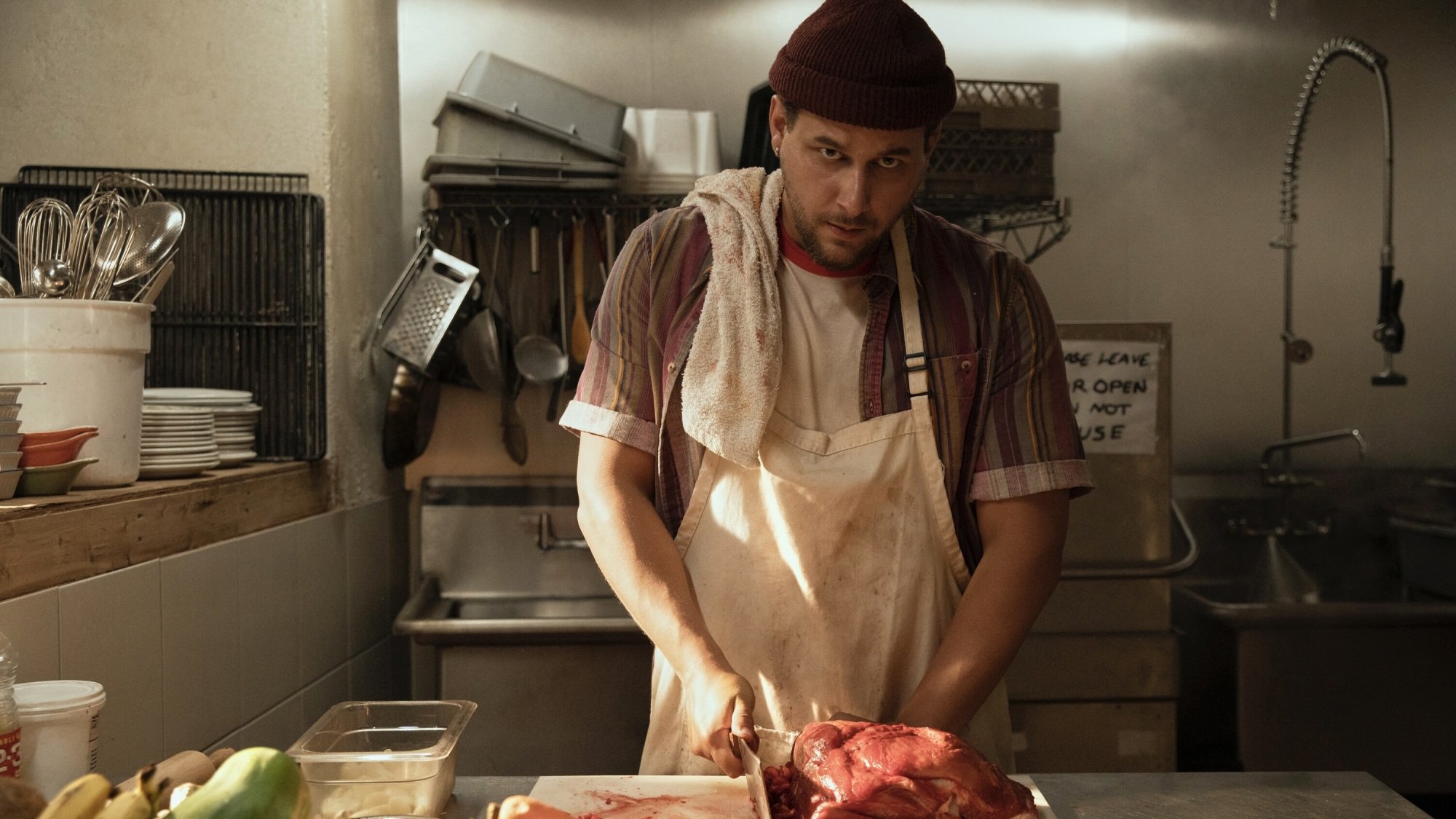 A man in an apron and maroon beanie chops meat with a cleaver in a restaurant kitchen.