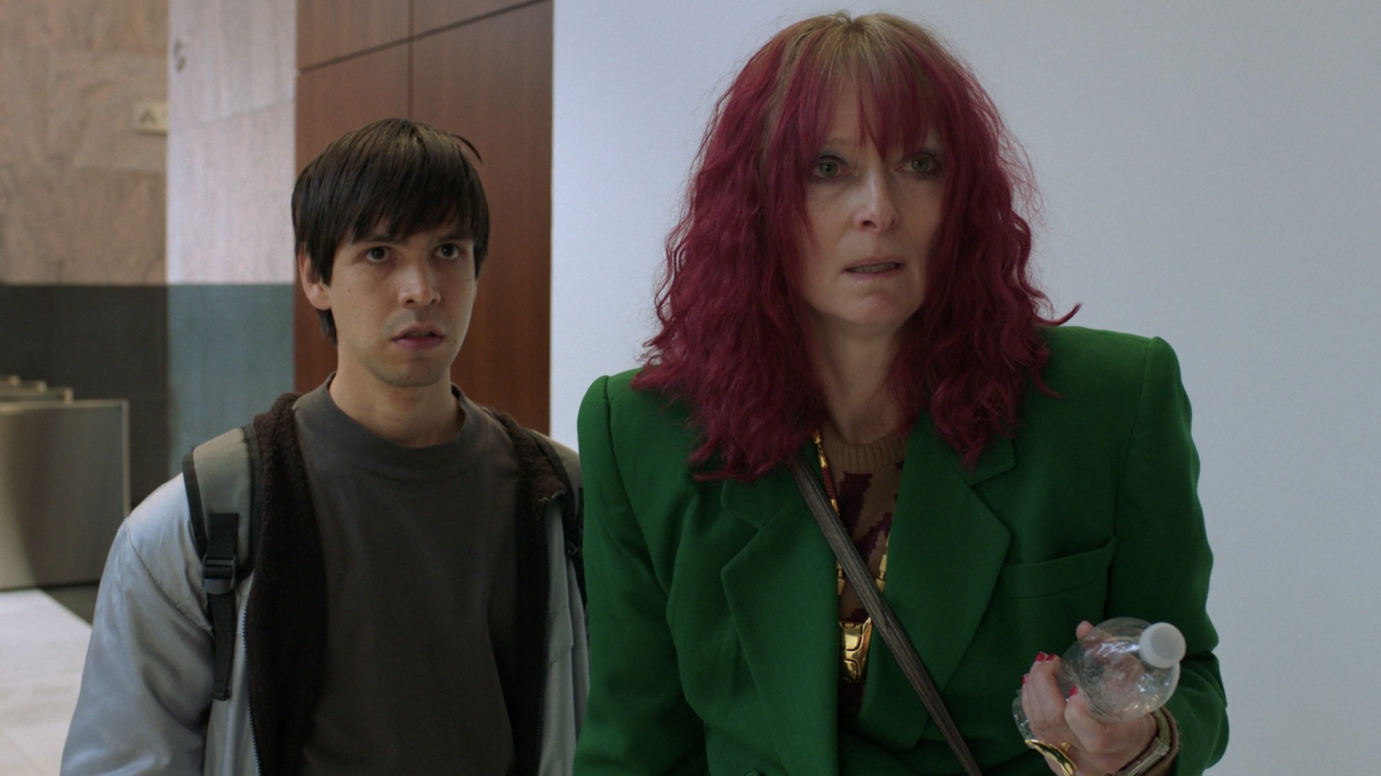 A man in a grey jacket and a woman with pink hair wearing a green jacket stand together with shocked looks on their faces.