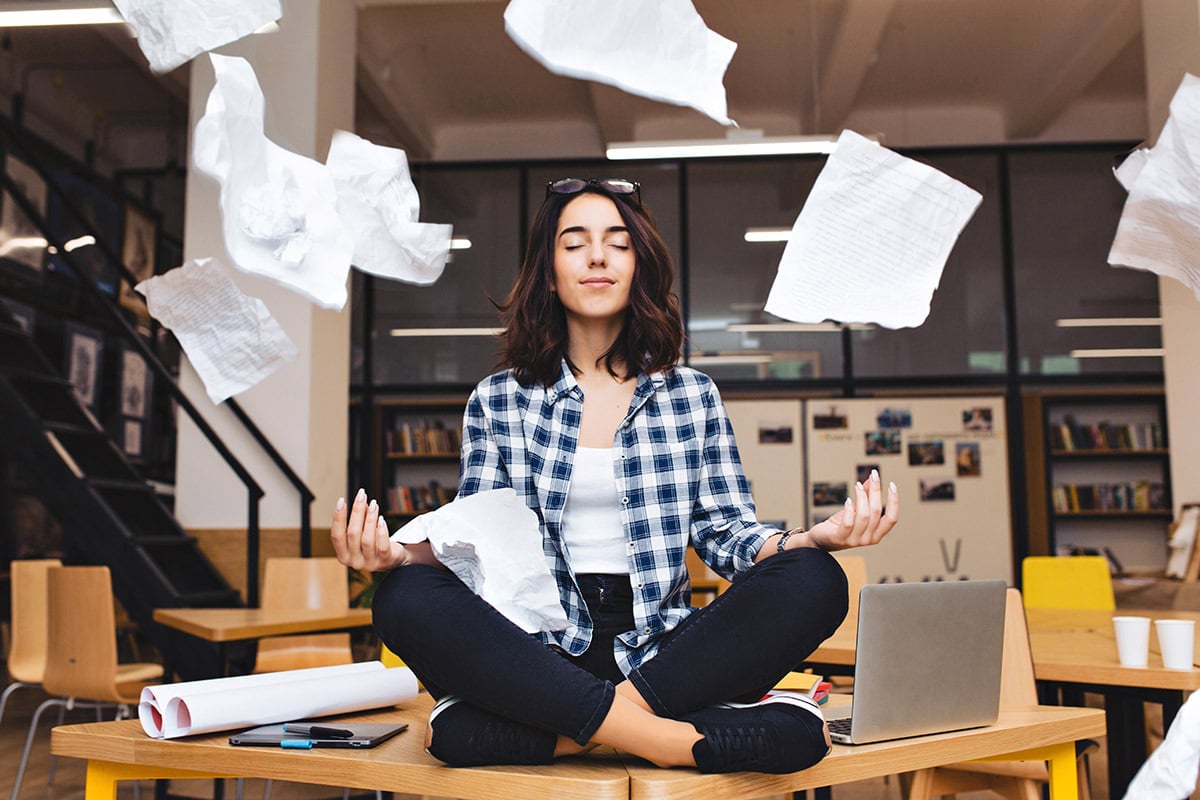 A woman sitting in lotus pose amidst a bunch of flying papers in an office. 
