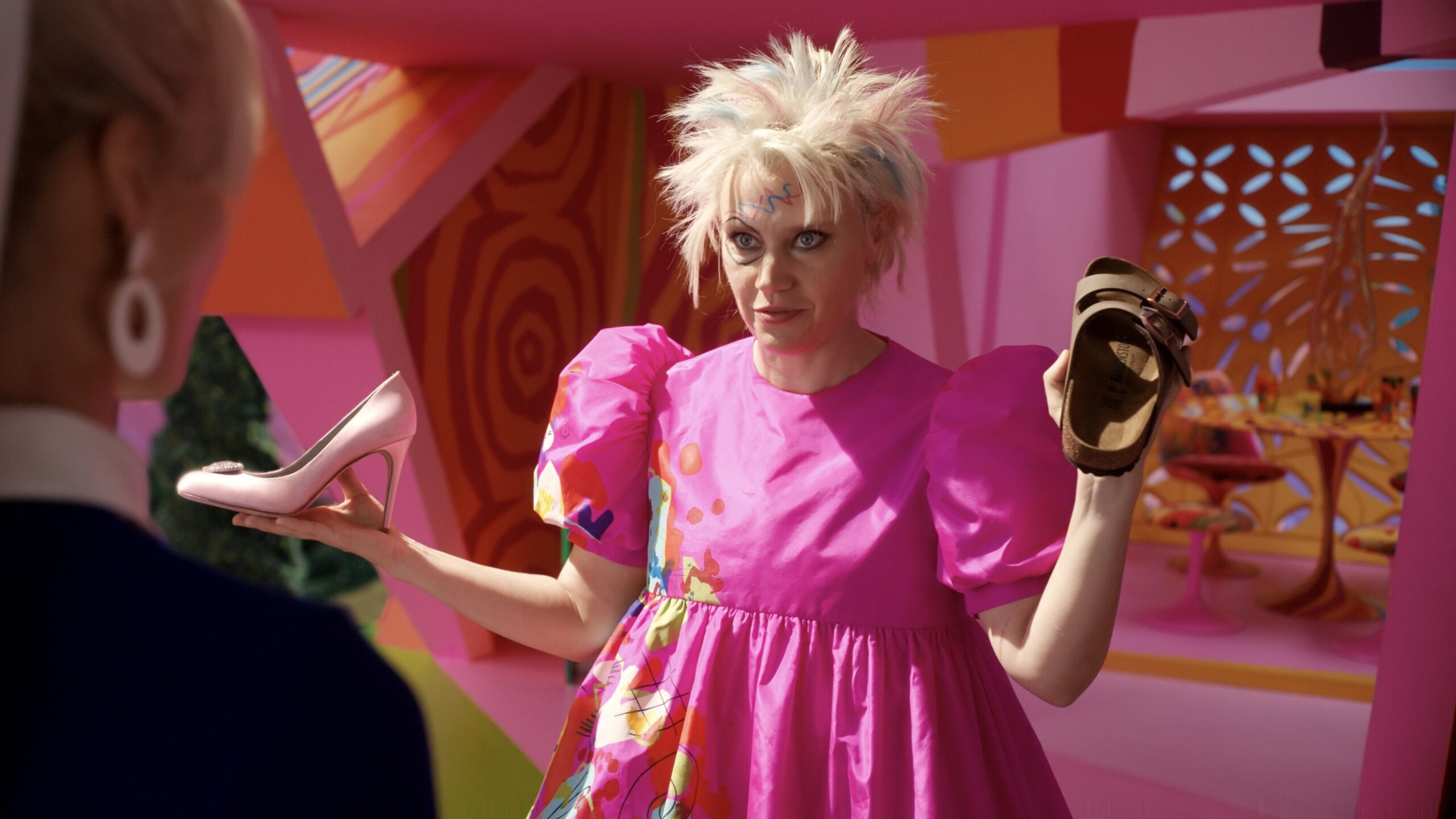 A woman in a pink dress with spiky blonde hair and crayon drawings all over her face holds a high heel in one hand and a Birkenstock sandal in the other.