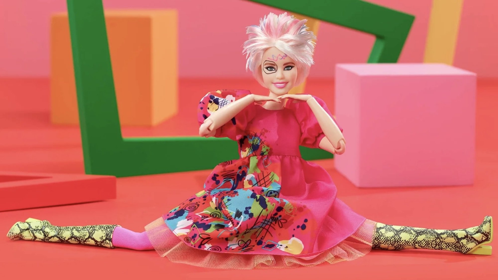 A Barbie doll in the splits; she has choppy, short blonde hair, a pink dress, green snakeskin boots, and crayon drawings all over here face.
