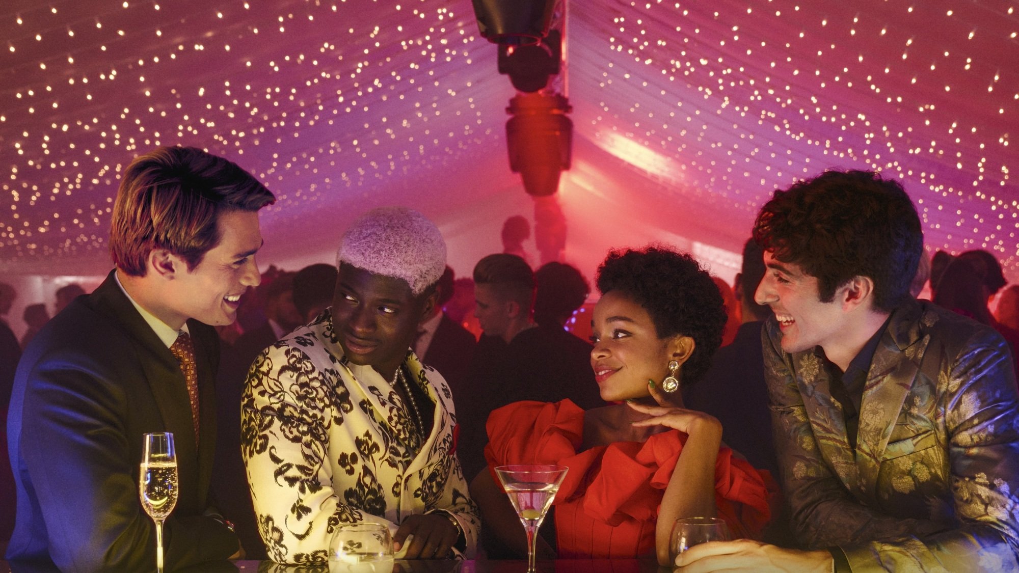 A group of four lavishly dressed men and woman hang out at a bar under a tent lit with red lights; a party rages behind them.