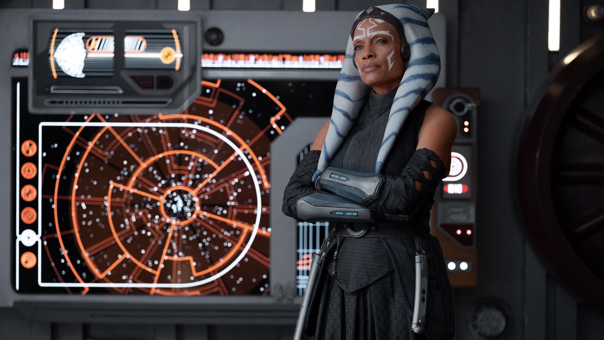 Ahsoka Tano stands with her arms crossed in front of a map lit up in orange.