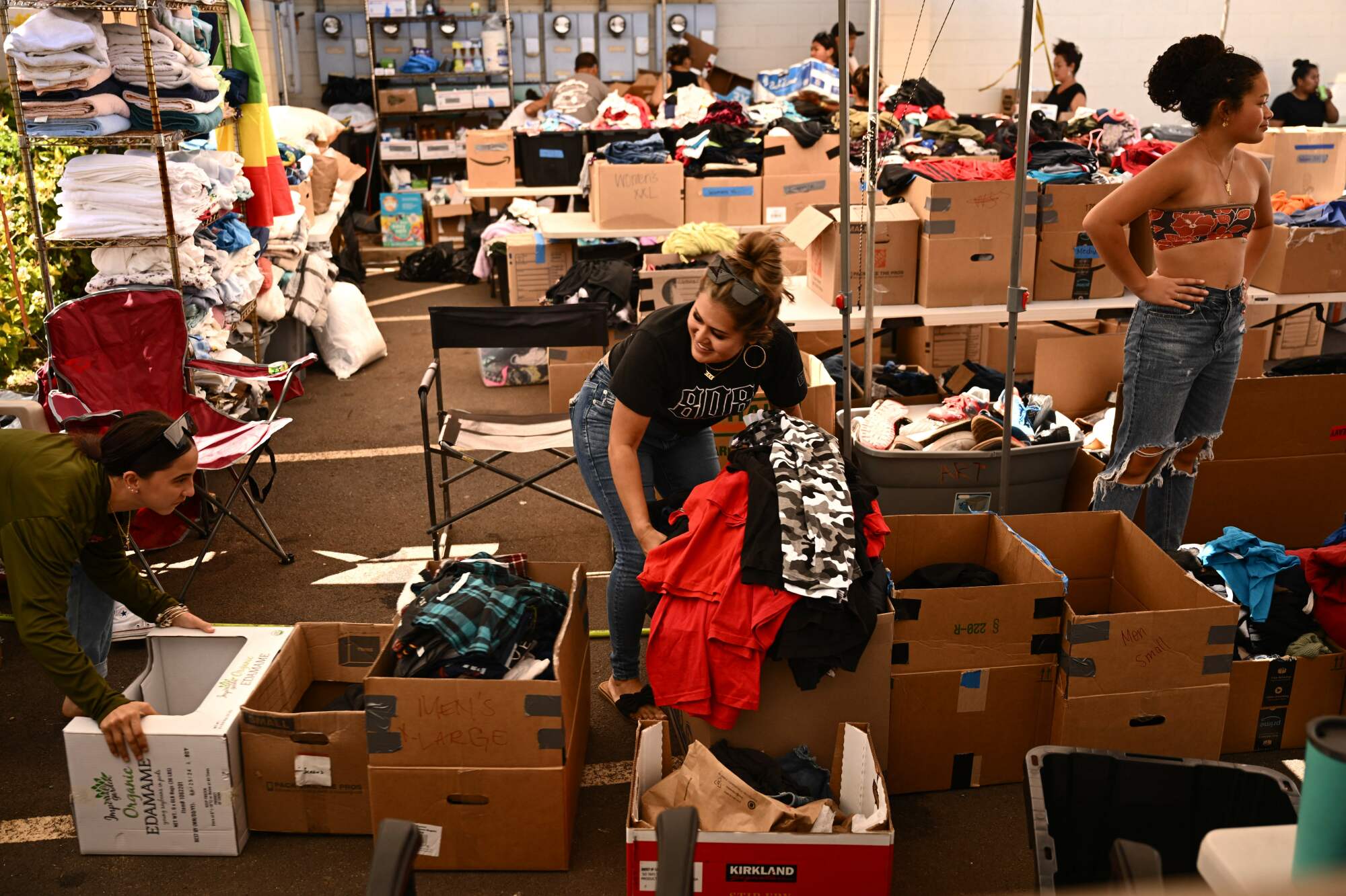 A woman bends over a brown box to pull out clothing, Behind her, brown boxes with goods fill a warehouse space.