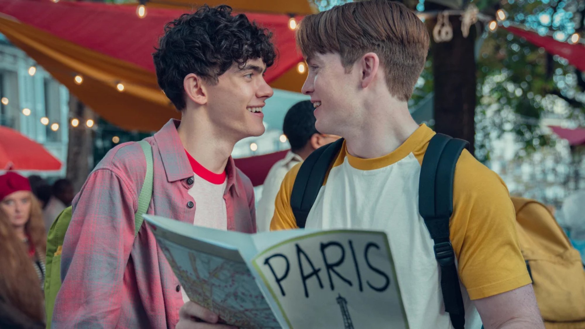 Two teenage boys look at each other lovingly while holding a tourist brochure about Paris.