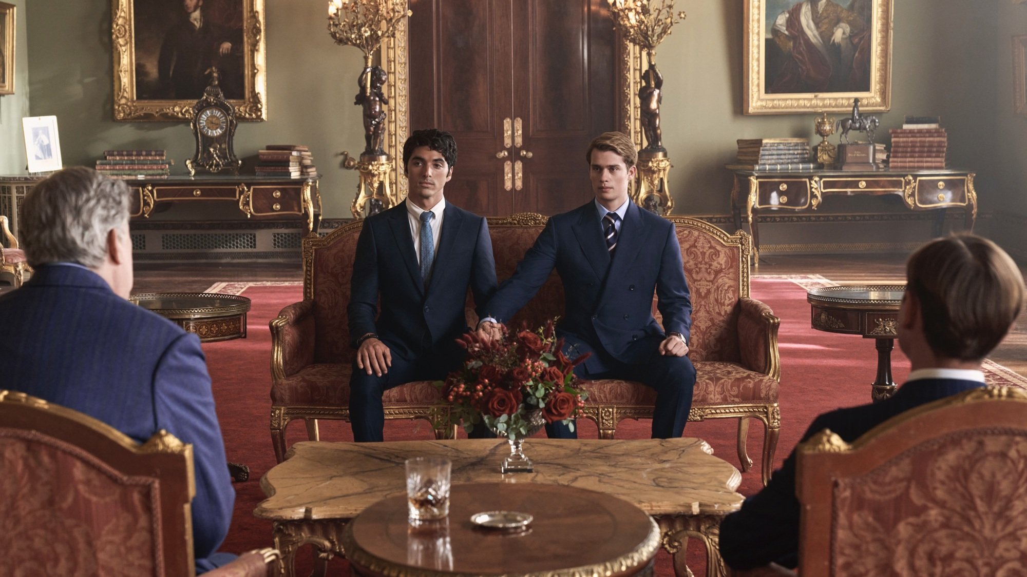 Two men in suits hold hands while seated on a red couch in Buckingham Palace.
