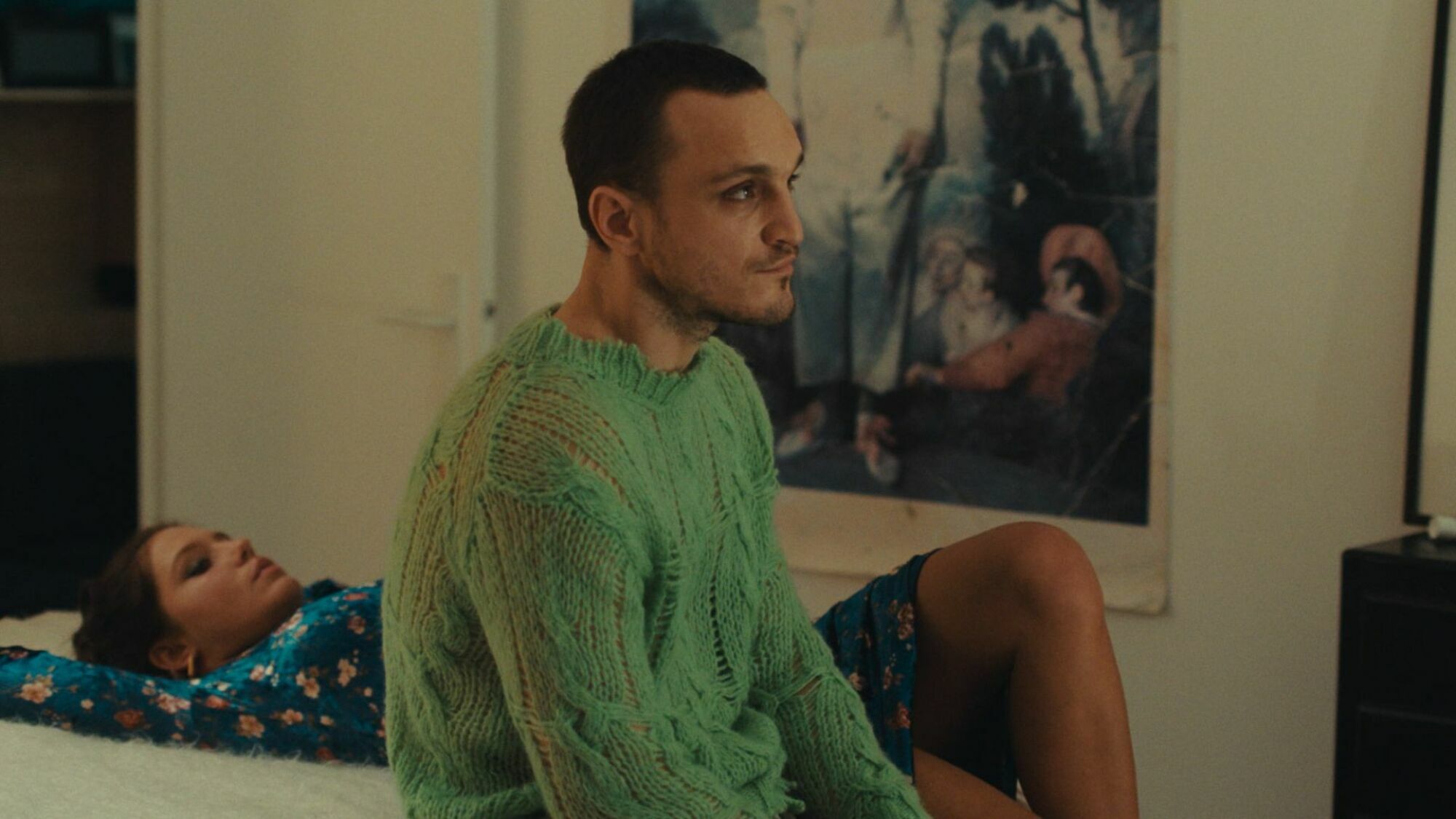 A man wearing a green sweater sits on a bed with a woman lying beside him. 