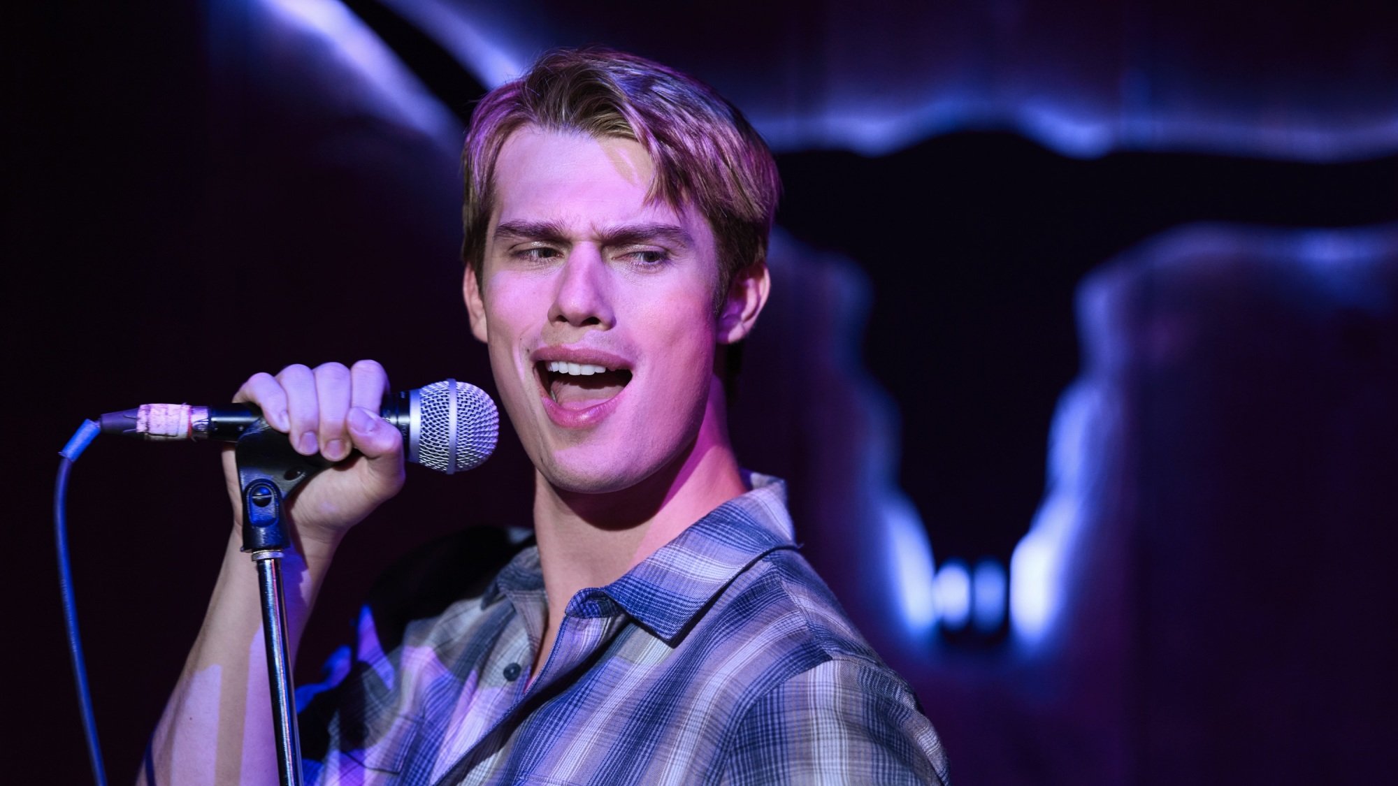 A blonde man in a blue checkered shirt sings passionate karaoke in a purple-lit bar.