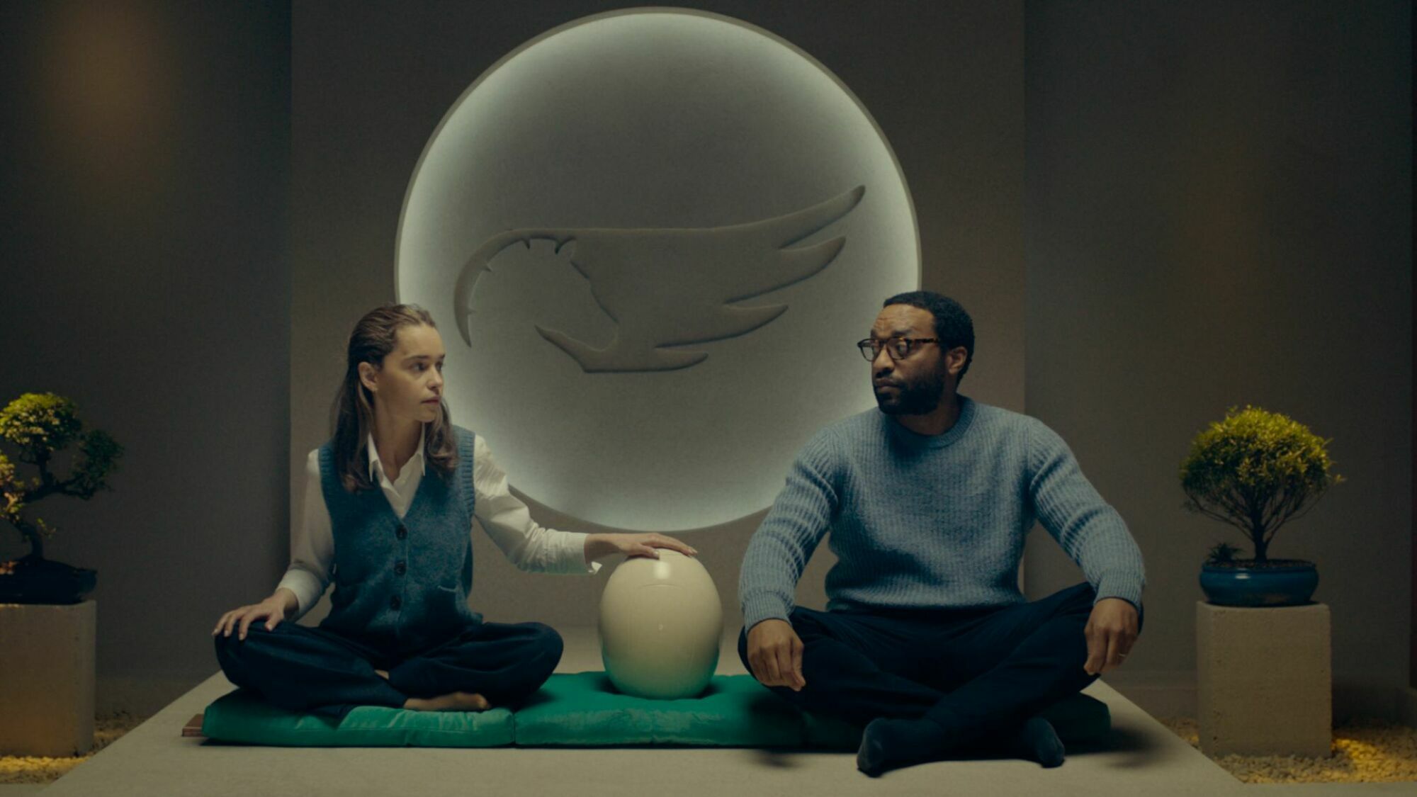 A man and a woman sit in a yoga room with a large egg between them. 