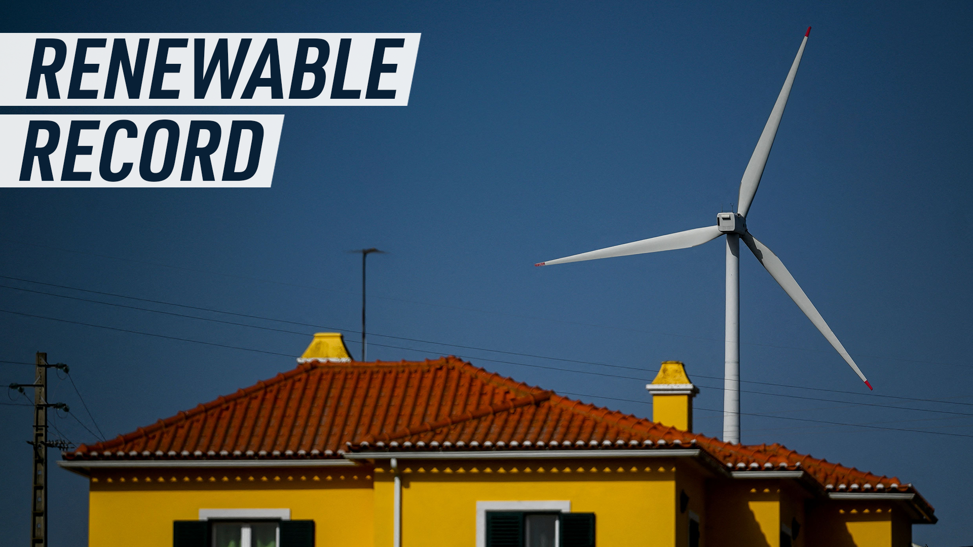 A picture shows the top of a typical portuguese house, yellow walls, dark green shutters, and a red rooftop. Behind it, against the blue sky a win turbine is peeking in the background.